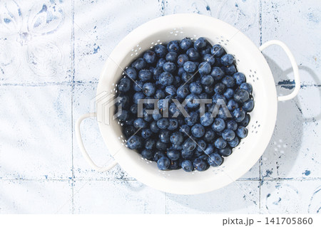 Fresh blueberries in a white colander placed on a light blue textured surface 141705860