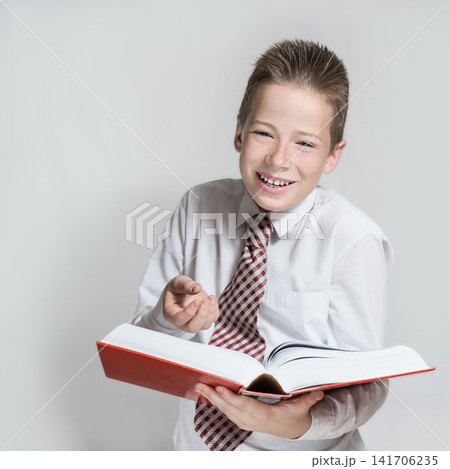 The nice smiling boy teenager in a white shirt and a tie reads a big red funny book on a gray background The nice smiling boy teenager in a white shirt and a tie reads a big red funny book on a gray background 141706235