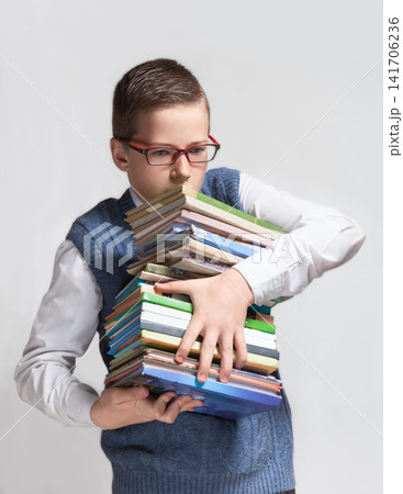 The cute smart schoolboy teenager in a glasses bears a heavy stack of books. The cute smart schoolboy teenager in a glasses bears a heavy stack of books. 141706236