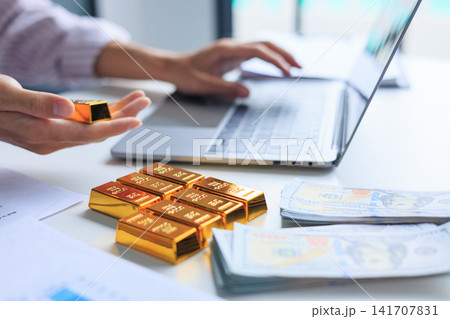 Investor Using Laptop to Manage Online Investment Trading Account with Gold Bars and Cash on the Table. Hedging Inflation with Precious Metals and Wealth Accumulation Concept. 141707831
