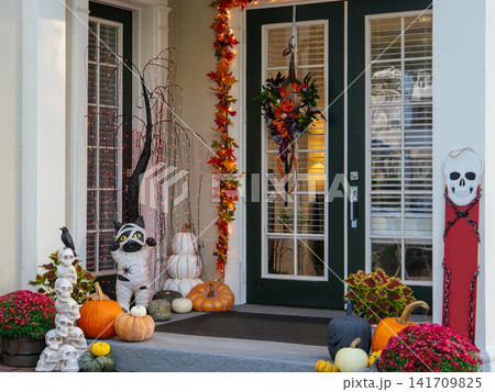 A front door is surrounded by pumpkins, a Halloween wreath, autumn leaves, and illuminated seasonal decorations. A festive display welcomes visitors with classic fall holiday decorations. 141709825