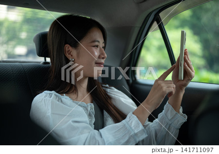 Young Asian woman taking a selfie photo with smartphone while sitting in back seat of a taxi cab car 141711097