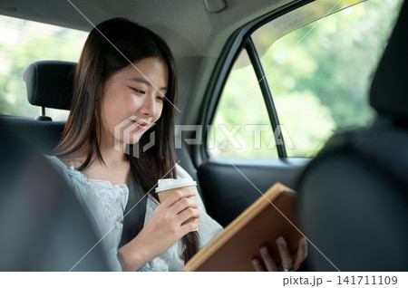 Young Asian woman holding coffee and reading a book while riding in the back seat of a taxi cab car. 141711109
