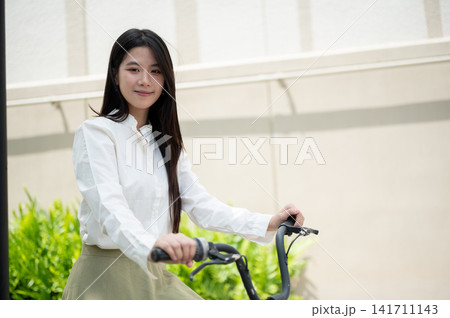 Young Asian woman riding a bicycle with friendly smile in urban streetscape under bright sunlight. Young Asian woman riding a bicycle with friendly smile in urban streetscape under bright sunlight. 141711143