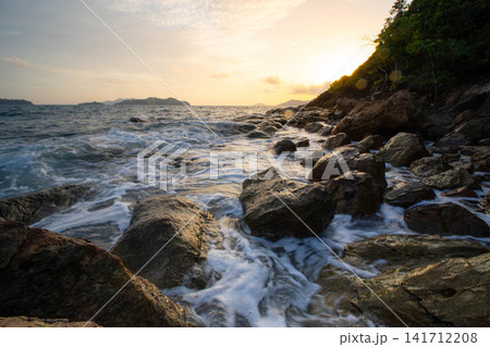 Ocean waves crashing on rocky shore with golden sunlight at sunset or sunrise 141712208