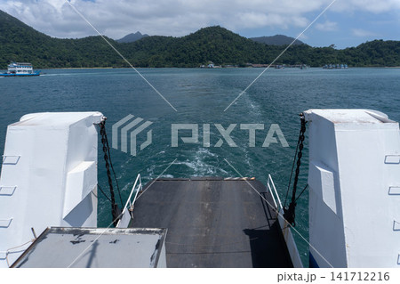 Ramp of a ferry boat navigating through deep blue sea towards island mountains under cloudy sky 141712216