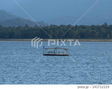 Small wooden raft floating on a vast blue lake with forest and mountain landscape Small wooden raft floating on a vast blue lake with forest and mountain landscape 141712229