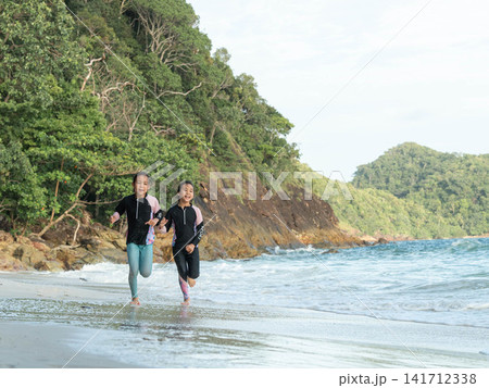 Two happy young Asian girls running together along the sandy beach near rocky cliff during summer trip Two happy young Asian girls running together along the sandy beach near rocky cliff during summer trip 141712338