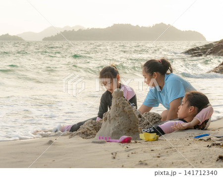Mother and daughters playing together covering legs with sand to create a mermaid tail shape on beach Mother and daughters playing together covering legs with sand to create a mermaid tail shape on beach 141712340