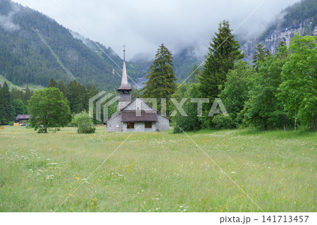 Beautiful walking trail in Kandersteg village near Oeschinen Lake. Traditional Swiss wooden chalet, church and summer wildflowers green meadow with stunning Swiss Alps mountains background Beautiful walking trail in Kandersteg village near Oeschinen Lake. Traditional Swiss wooden chalet, church and summer wildflowers green meadow with stunning Swiss Alps mountains background 141713457
