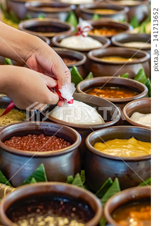 Traditional Indonesian dessert assortment with sweet porridge served in clay pots. Hands packing takeaway food in market setting, authentic Asian cuisine. 141713652