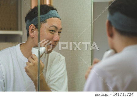 三面鏡の前で美顔器を頬に当てる40代男性のメンズスキンケア 三面鏡の前で美顔器を頬に当てる40代男性のメンズスキンケア 141713724