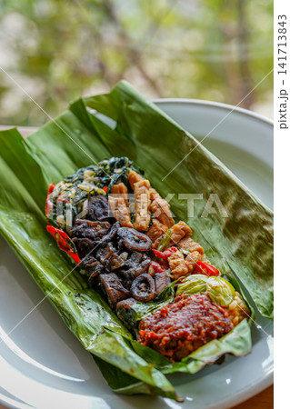 Authentic Indonesian banana leaf wrapped rice with black squid in squid ink, tempeh and sambal served on a white plate. Perfect for restaurant, culinary and travel concepts. Authentic Indonesian banana leaf wrapped rice with black squid in squid ink, tempeh and sambal served on a white plate. Perfect for restaurant, culinary and travel concepts. 141713843