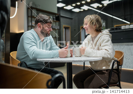 Middle aged man and young woman smiling across marble table in modern coffee shop interior, sharing relaxed daytime conversation with warm industrial lighting, with conversation, smiling, cafe Middle aged man and young woman smiling across marble table in modern coffee shop interior, sharing relaxed daytime conversation with warm industrial lighting, with conversation, smiling, cafe 141714413