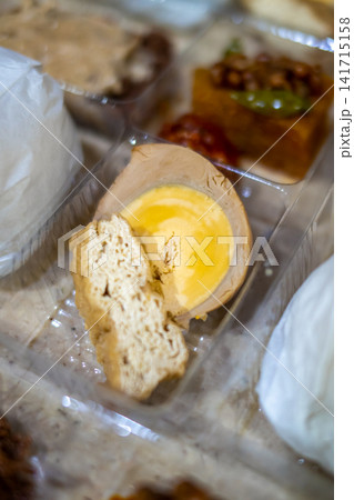 Close-up of an Indonesian braised egg with yellow yolk and crispy kerupuk served with traditional side dishes in a clear takeaway food container. Close-up of an Indonesian braised egg with yellow yolk and crispy kerupuk served with traditional side dishes in a clear takeaway food container. 141715158