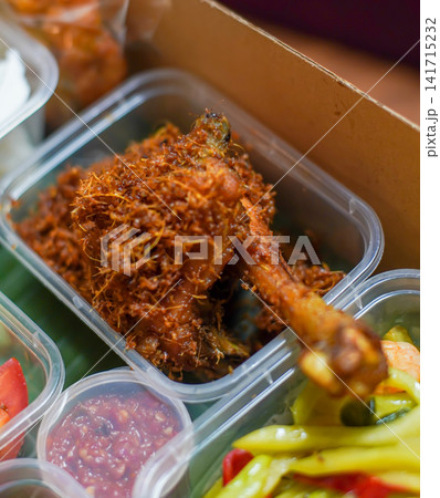 Crispy Indonesian fried chicken covered with fragrant fried galangal, served with spicy sambal and colorful pickled vegetables in takeaway containers, representing Southeast Asian cuisine. Crispy Indonesian fried chicken covered with fragrant fried galangal, served with spicy sambal and colorful pickled vegetables in takeaway containers, representing Southeast Asian cuisine. 141715232
