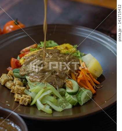 Indonesian gado gado with fresh vegetables, tofu, and peanut sauce pouring. Healthy Asian dish with authentic taste and colorful presentation. Indonesian gado gado with fresh vegetables, tofu, and peanut sauce pouring. Healthy Asian dish with authentic taste and colorful presentation. 141715412