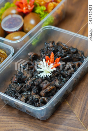Traditional Indonesian cumi hitam, squid cooked in rich black ink sauce with spices, served in plastic container, exotic Asian seafood meal close up Traditional Indonesian cumi hitam, squid cooked in rich black ink sauce with spices, served in plastic container, exotic Asian seafood meal close up 141715421