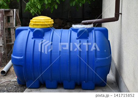 Large water storage tank is positioned beside a building wall, connected to a downspout pipe for collecting rainwater. System for rainwater harvesting, resource management, eco-friendly living Large water storage tank is positioned beside a building wall, connected to a downspout pipe for collecting rainwater. System for rainwater harvesting, resource management, eco-friendly living 141715852