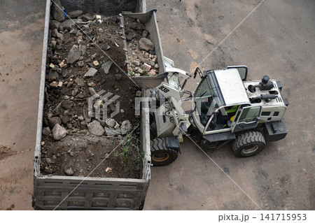 Aerial view of construction site where a front loader is loading soil, rocks, and debris into a dump truck, industrial work, excavation, and earthmoving processes Aerial view of construction site where a front loader is loading soil, rocks, and debris into a dump truck, industrial work, excavation, and earthmoving processes 141715953