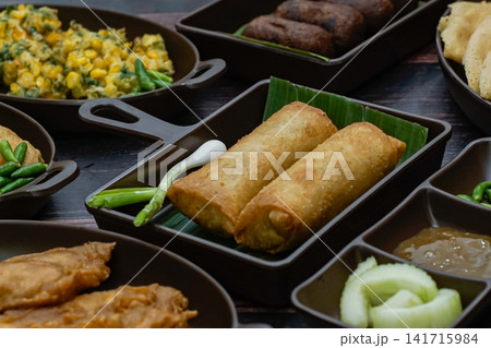 Traditional Indonesian lumpia fried spring rolls served on banana leaf with green chilies and surrounded by assorted local dishes, showcasing Indonesian food culture. 141715984