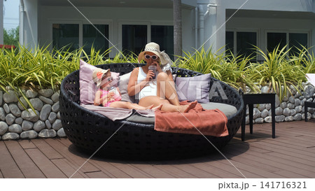 Mother in white swimsuit and straw hat with little daughter in pink swimsuit and straw hat both drinking fruit cocktails in comfortable chairs beside a resort pool. Family leisure concept Mother in white swimsuit and straw hat with little daughter in pink swimsuit and straw hat both drinking fruit cocktails in comfortable chairs beside a resort pool. Family leisure concept 141716321