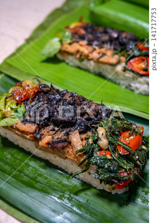 Traditional Indonesian seafood rice with black squid ink, vegetables, herbs, and sambal served on banana leaf. Authentic Southeast Asian cuisine suitable for travel, restaurant, and food publications. Traditional Indonesian seafood rice with black squid ink, vegetables, herbs, and sambal served on banana leaf. Authentic Southeast Asian cuisine suitable for travel, restaurant, and food publications. 141717753