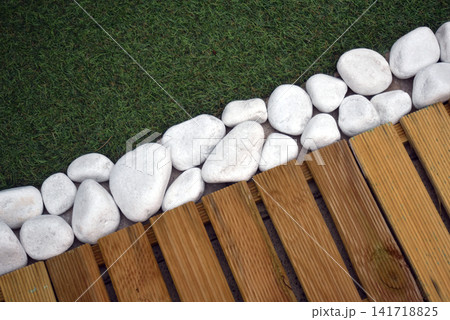 Detail of a garden pathway featuring artificial grass, wooden decking tiles and white pebbles along the border, creating a modern outdoor landscaping design. 141718825
