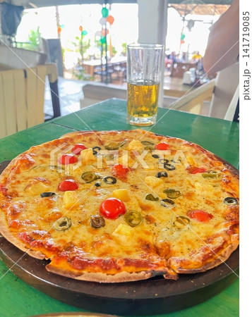 木製ボードに載るプチトマトやパイナップルがトッピングされた大皿ピザとグラスの飲み物 141719085