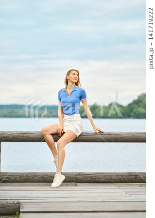 Woman in blue top and white shorts sitting on wooden dock near lake during cloudy day in summer Woman in blue top and white shorts sitting on wooden dock near lake during cloudy day in summer 141719222