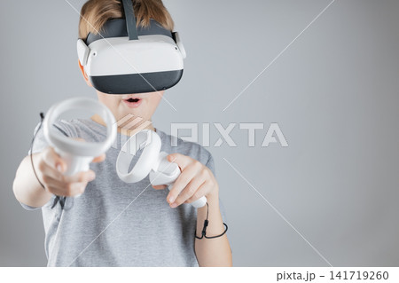 Excited Young Boy in Gray Shirt Immersed in Virtual Reality Gaming with White VR Headset and Game Controllers on Gray Background Excited Young Boy in Gray Shirt Immersed in Virtual Reality Gaming with White VR Headset and Game Controllers on Gray Background 141719260