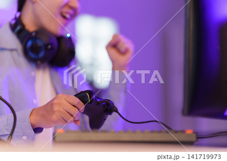Asian gamer holding controller celebrating video game victory in purple room Asian gamer holding controller celebrating video game victory in purple room 141719973