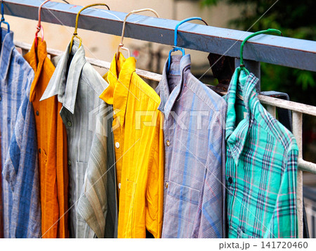 Colorful shirts hanging for sale at a market stall in Myanmar, showing everyday street local trade and local textiles. Open-air market rows of colorful shirts hanging on a railing. 141720460