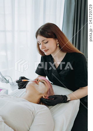 professional aesthetician applies a creamy face mask on young woman face using a brush professional aesthetician applies a creamy face mask on young woman face using a brush 141720729