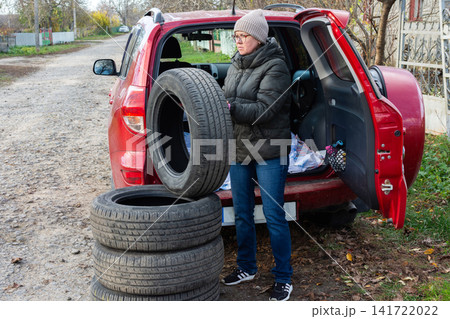 A person is swapping summer tires for winter tires near a red car on a gravel road. This activity takes place during the fall season as the leaves change 141722022