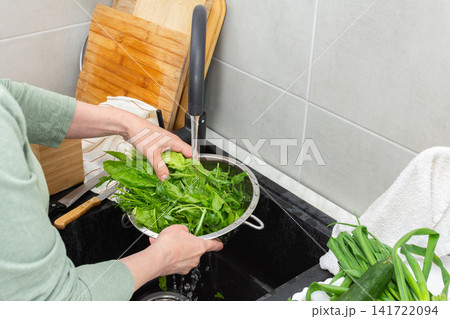 Hands are holding a colander filled with fresh organic local vegetables under running water in a kitchen sink. This is done to clean the produce before cooking 141722094