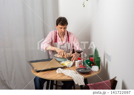 A 53-year-old man is preparing chicken for pre-Christmas dinner. He seasons the chicken in a bowl. Nearby are potatoes and a glass of wine on the table 141722097