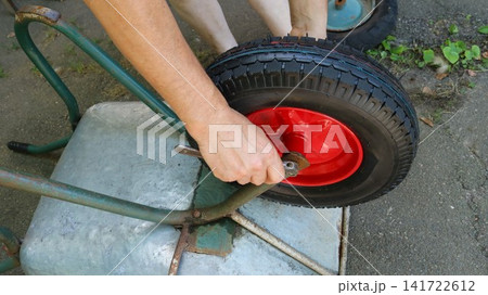 A close-up view of a garden wheelbarrow being repaired. A man's hands use a wrench to tighten a new wheel with a bright red rim and a black tire, mounted on a metal frame. 141722612