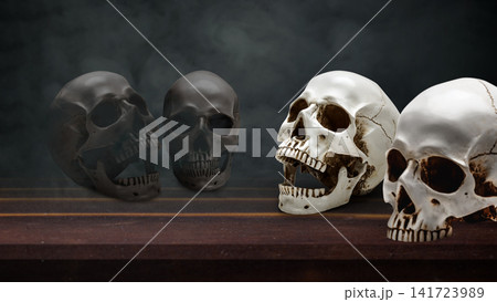 Four human skulls, two black and two white, arranged on a dark wooden table against a moody, textured background, representing themes of life, death, duality, and mortality. Concept of Halloween Four human skulls, two black and two white, arranged on a dark wooden table against a moody, textured background, representing themes of life, death, duality, and mortality. Concept of Halloween 141723989