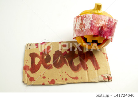 Close-up of a small Halloween pumpkin wrapped in a red-stained bandage next to a piece of cardboard with the word Death written in a bloody font against a plain white background. Concept of Halloween Close-up of a small Halloween pumpkin wrapped in a red-stained bandage next to a piece of cardboard with the word Death written in a bloody font against a plain white background. Concept of Halloween 141724040