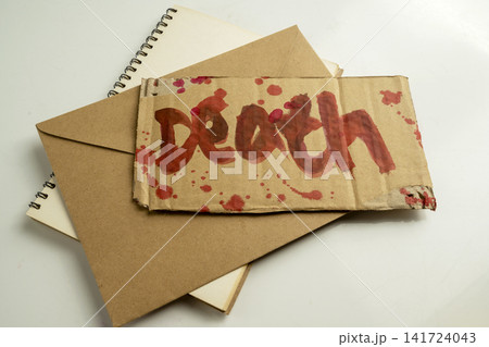 A piece of cardboard with the word death written in red blood, placed on top of a brown paper envelope and a spiral notebook, creating a dark, suspenseful concept. Concept of Halloween A piece of cardboard with the word death written in red blood, placed on top of a brown paper envelope and a spiral notebook, creating a dark, suspenseful concept. Concept of Halloween 141724043