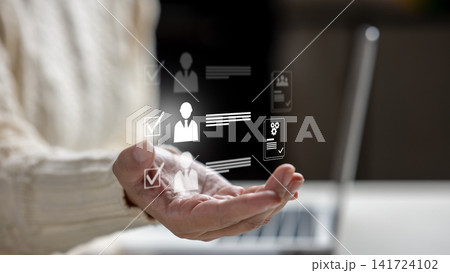Female hand holding virtual HR icons representing recruitment, candidate selection, and user profiles with a laptop in the background. HR Recruitment concept Female hand holding virtual HR icons representing recruitment, candidate selection, and user profiles with a laptop in the background. HR Recruitment concept 141724102