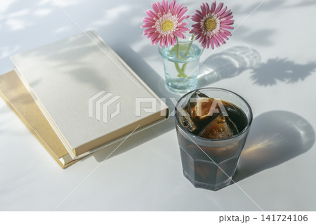 アイスコーヒーとピンクのガーベラ、白いハードカバーの本が並ぶ夏の読書イメージ アイスコーヒーとピンクのガーベラ、白いハードカバーの本が並ぶ夏の読書イメージ 141724106