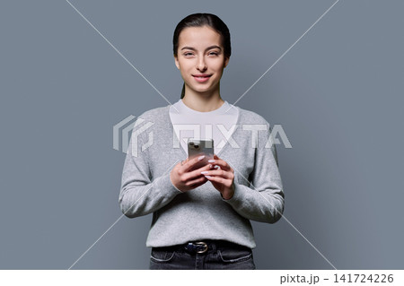 Smiling young woman holding smartphone and looking at camera over grey background Smiling young woman holding smartphone and looking at camera over grey background 141724226
