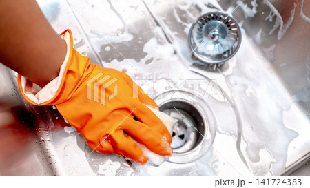 Housekeeper hand with orange rubber glove scrubbing on kitchen sink drainer Housekeeper hand with orange rubber glove scrubbing on kitchen sink drainer 141724383