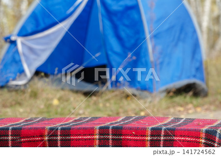 Cozy outdoor camping setup featuring a red plaid blanket with a blue tent in soft autumn light Cozy outdoor camping setup featuring a red plaid blanket with a blue tent in soft autumn light 141724562