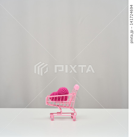 A pink shopping cart with a knitted pink heart inside stands on a white table against a light background 141724694