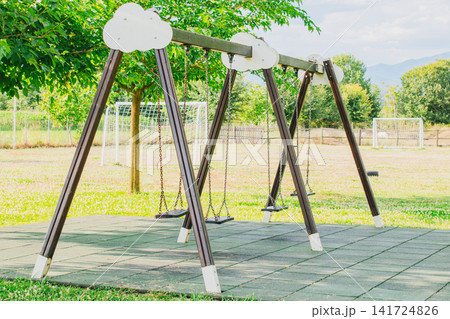 Empty wooden swing set with chain seats standing on soft rubber safety tiles at outdoor public kids playground with green trees and lawn background on sunny summer day Empty wooden swing set with chain seats standing on soft rubber safety tiles at outdoor public kids playground with green trees and lawn background on sunny summer day 141724826