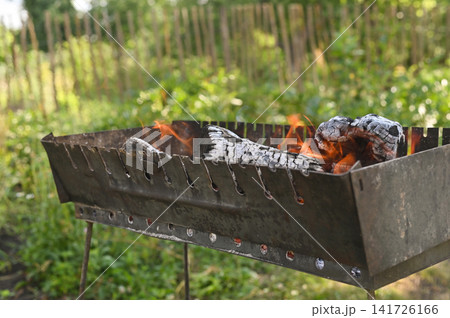 Burning firewood with orange flames inside metal grill outdoors. Firewood creates hot charcoal for summer barbecue picnic. Natural birch firewood glows in brazier on blurred background. Burning firewood with orange flames inside metal grill outdoors. Firewood creates hot charcoal for summer barbecue picnic. Natural birch firewood glows in brazier on blurred background. 141726166