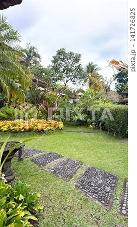 Lush tropical garden path creating a tranquil ambiance, displaying vibrant green foliage, exotic plants, and traditional stone stepping stones on green grass Lush tropical garden path creating a tranquil ambiance, displaying vibrant green foliage, exotic plants, and traditional stone stepping stones on green grass 141726825